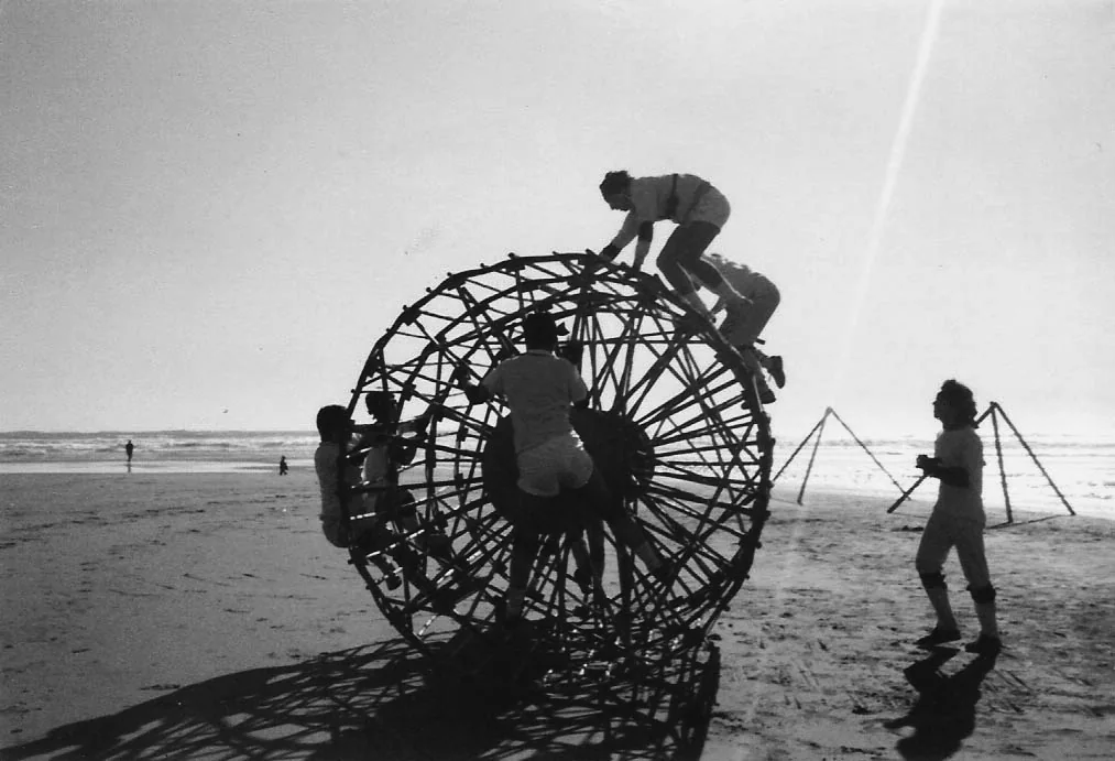 People climbing a large circular experimental structure on a beach.